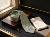 The Highland Thistle Silk Tie coiled on dark walnut with a pocket square and brass collar bar