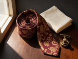 The Buckingham Wine Floral Silk Tie coiled on dark walnut with a pocket square and brass collar bar