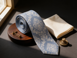 The Ascot Fleur Silk Tie coiled on dark walnut with a pocket square and brass collar bar