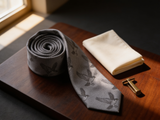 The Balmoral Falcon Silk Tie coiled on dark walnut with a pocket square and brass collar bar