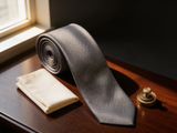 The Mayfair Lattice Silk Tie coiled on dark walnut with a pocket square and brass collar bar