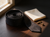 The Mayfair Charcoal Diamond Tie coiled on dark walnut with a pocket square and brass collar bar