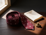 The Mayfair Burgundy Diamond Tie coiled on dark walnut with a pocket square and brass collar bar