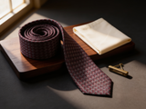 The Westminster Burgundy Basketweave Tie coiled on dark walnut with a pocket square and brass collar bar