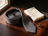 The Bond Textured Grenadine Tie coiled on dark walnut with a pocket square and brass collar bar