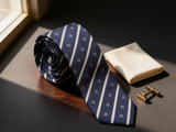 The Mayfair Club Stripe Tie coiled on dark walnut with a pocket square and brass collar bar