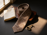 The Belgravia Taupe Paisley Tie coiled on dark walnut with a pocket square and brass collar bar