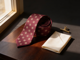 Sovereign Burgundy Rosette Tie coiled on dark walnut with a pocket square and brass collar bar